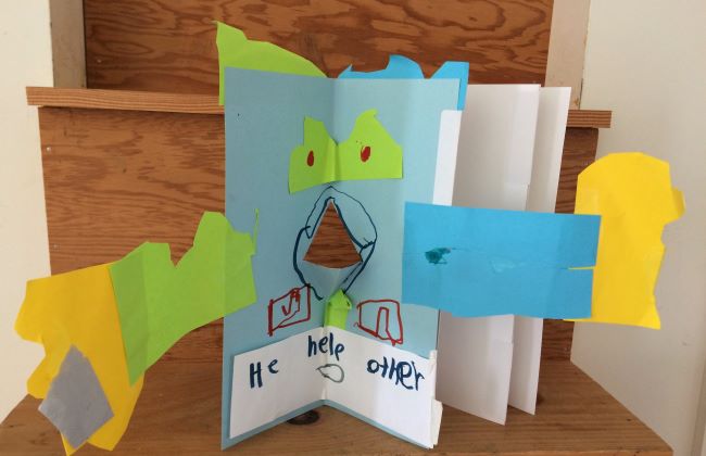 A child's book with a figure that has a pop up nose and long arms made from paper. The text says, "He help other."
